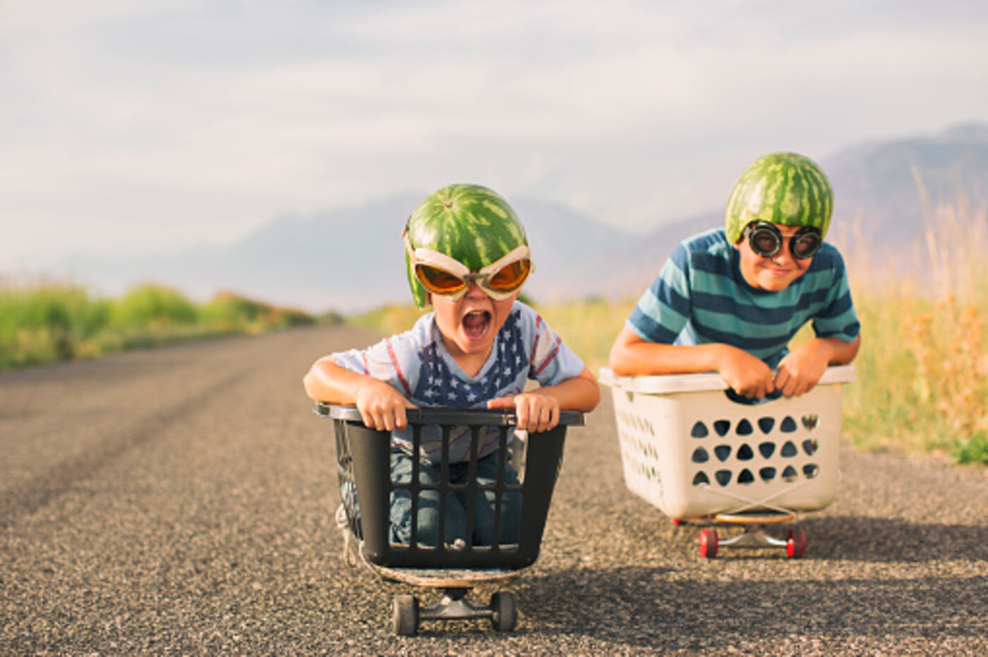 Two children in goggles racing laundry baskets down a road